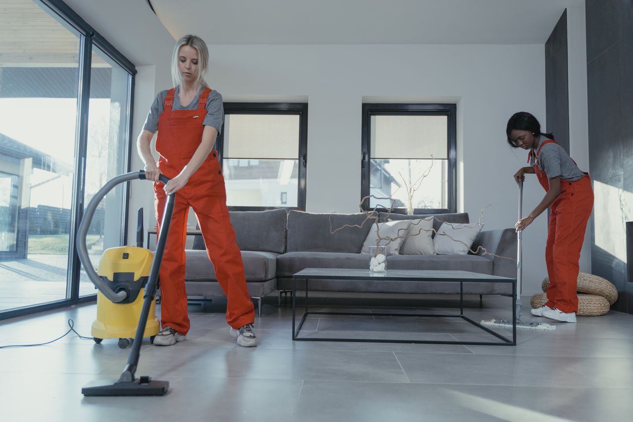 home-img Two women in red overalls vacuuming and sweeping a modern living room.